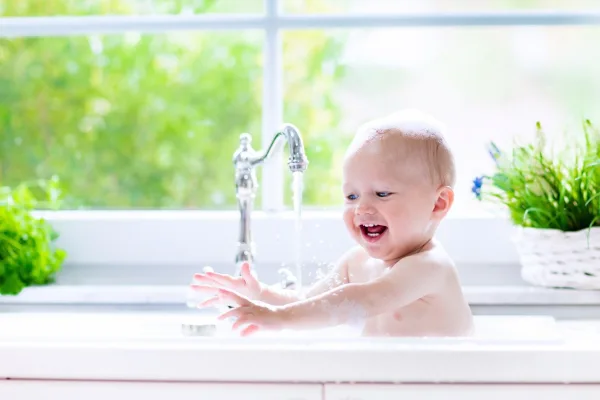 Smiling baby splashing in clean running water at a bright kitchen sink