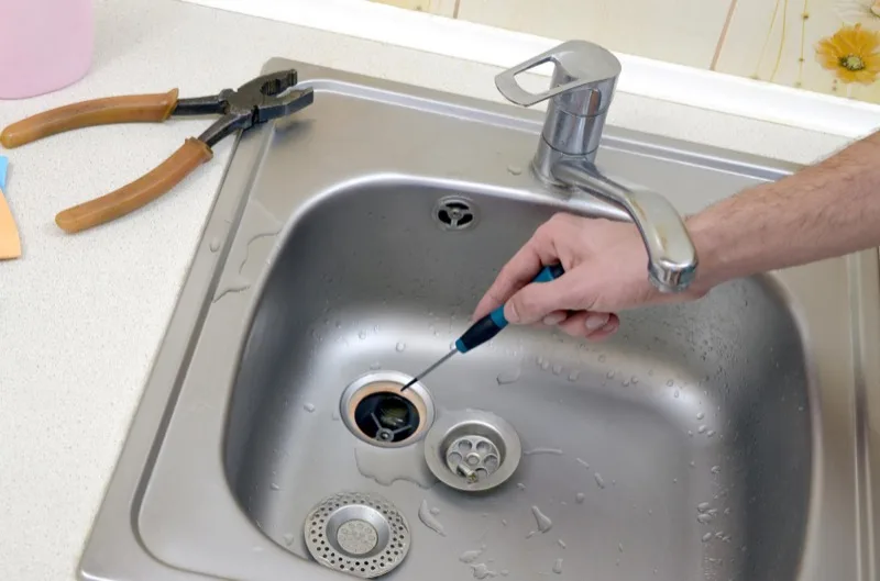 Hand working a tool into a stainless steel kitchen sink drain with pliers resting on the counter