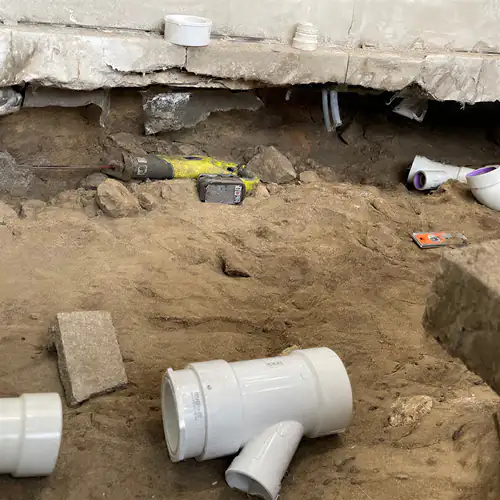 Excavation under a cut concrete slab showing the old dark cast iron drain pipe and new white PVC fittings waiting in the sand