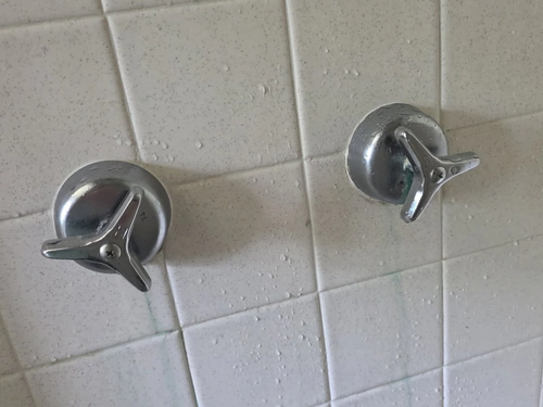 Two chrome cross handles on separate escutcheons in a white square tile shower wall