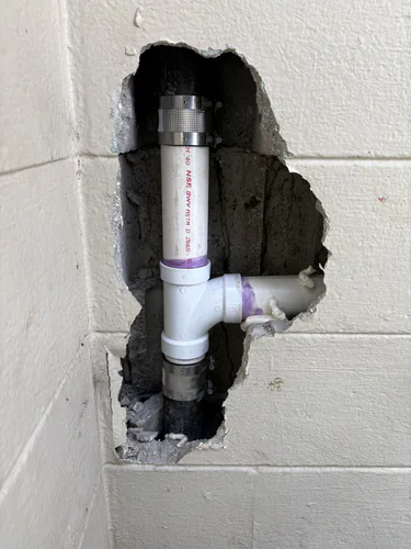 New white Schedule 40 PVC sanitary tee joined to an old cast iron drain stack with stainless shielded couplings inside an opened block wall in downtown Orlando