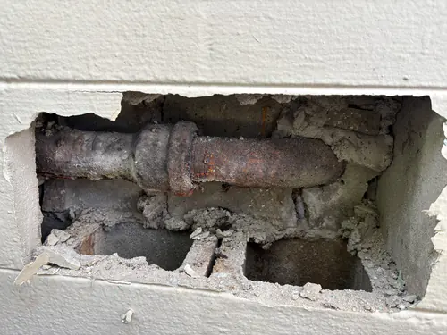 Before the repair: a rusted horizontal cast iron drain pipe with a hub joint, exposed through a rough opening in a painted concrete block wall